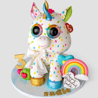A colorful unicorn cake decorated with rainbow spots, large eyes, a gold horn, and ears. It has a gold “3,” a rainbow, a heart, and a “Happy Birthday Sadie” message on a white cake board.