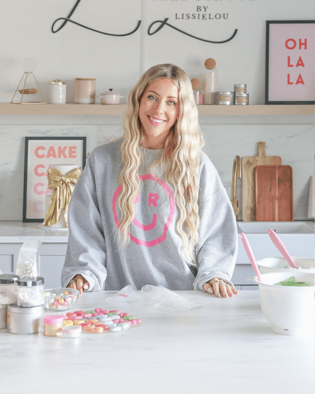 A smiling woman stands in a bright kitchen, wearing a grey Jenna Rae Cakes sweatshirt with a pink smiley face. Colorful baking decorations and jars surround her as she promotes Jenna Rae Cakes’ Online Classes.