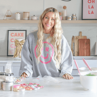 A smiling woman stands in a bright kitchen, wearing a grey Jenna Rae Cakes sweatshirt with a pink smiley face. Colorful baking decorations and jars surround her as she promotes Jenna Rae Cakes’ Online Classes.