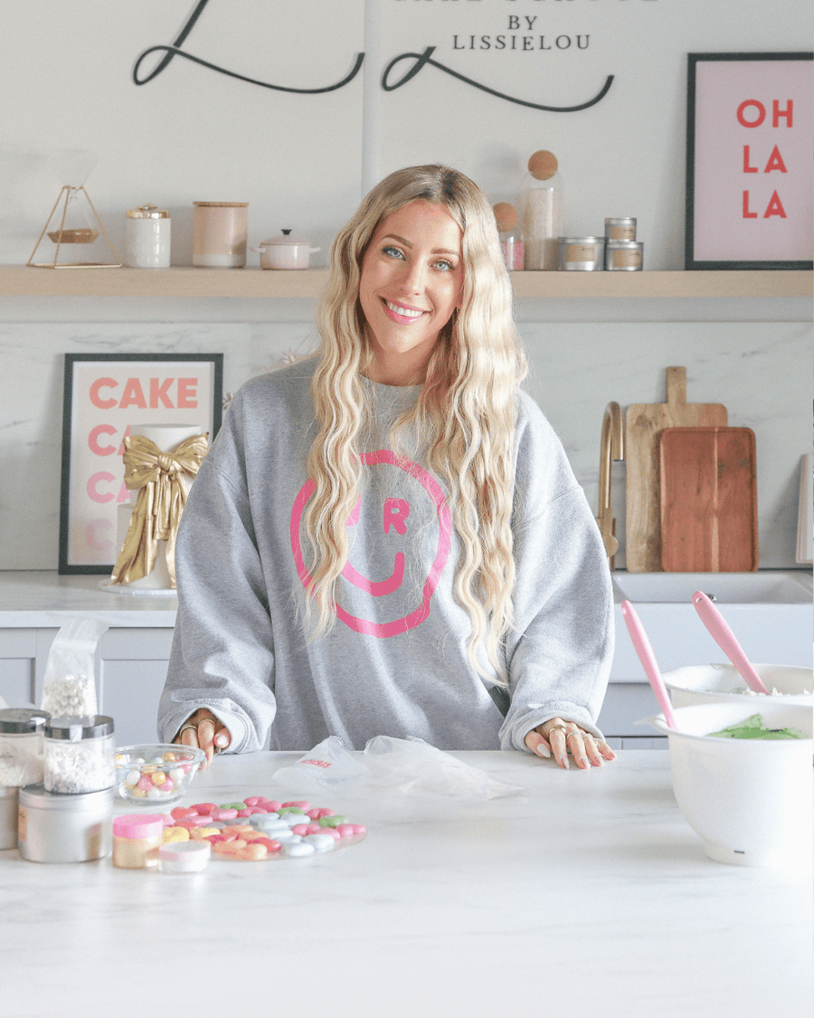 A smiling woman stands in a bright kitchen, wearing a grey Jenna Rae Cakes sweatshirt with a pink smiley face. Colorful baking decorations and jars surround her as she promotes Jenna Rae Cakes’ Online Classes.