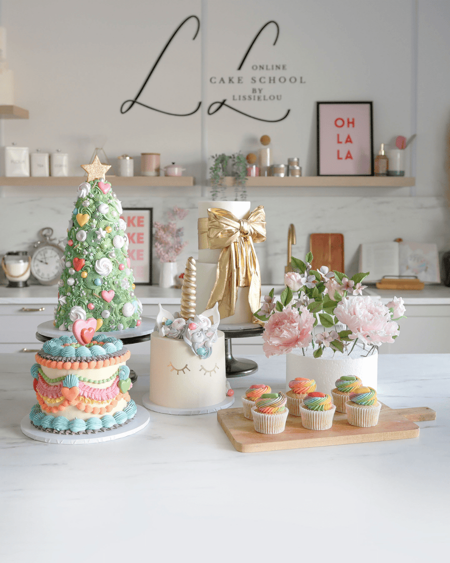 A vibrant kitchen display showcases Jenna Rae Cakes Online Classes with cakes like a unicorn cake, pink floral cake, Christmas tree cake, and pastel cupcakes on a marble counter, accented by flowers and contemporary decor.