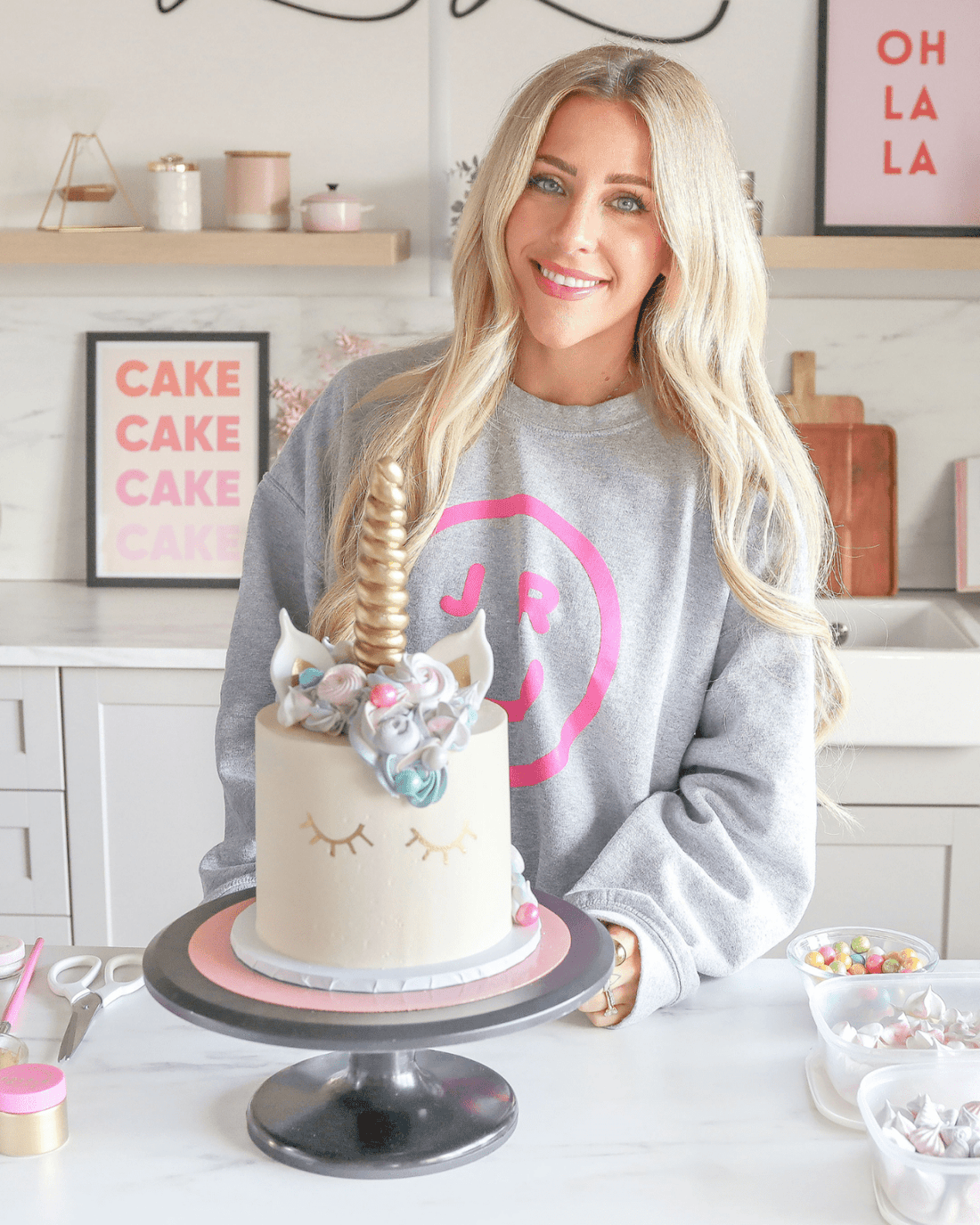 A smiling woman in a gray sweatshirt with a pink circle logo stands in a bright kitchen behind a unicorn cake. Baking supplies are on the counter. Jenna Rae Cakes offers Online Classes to help you create magical cakes like this at home.