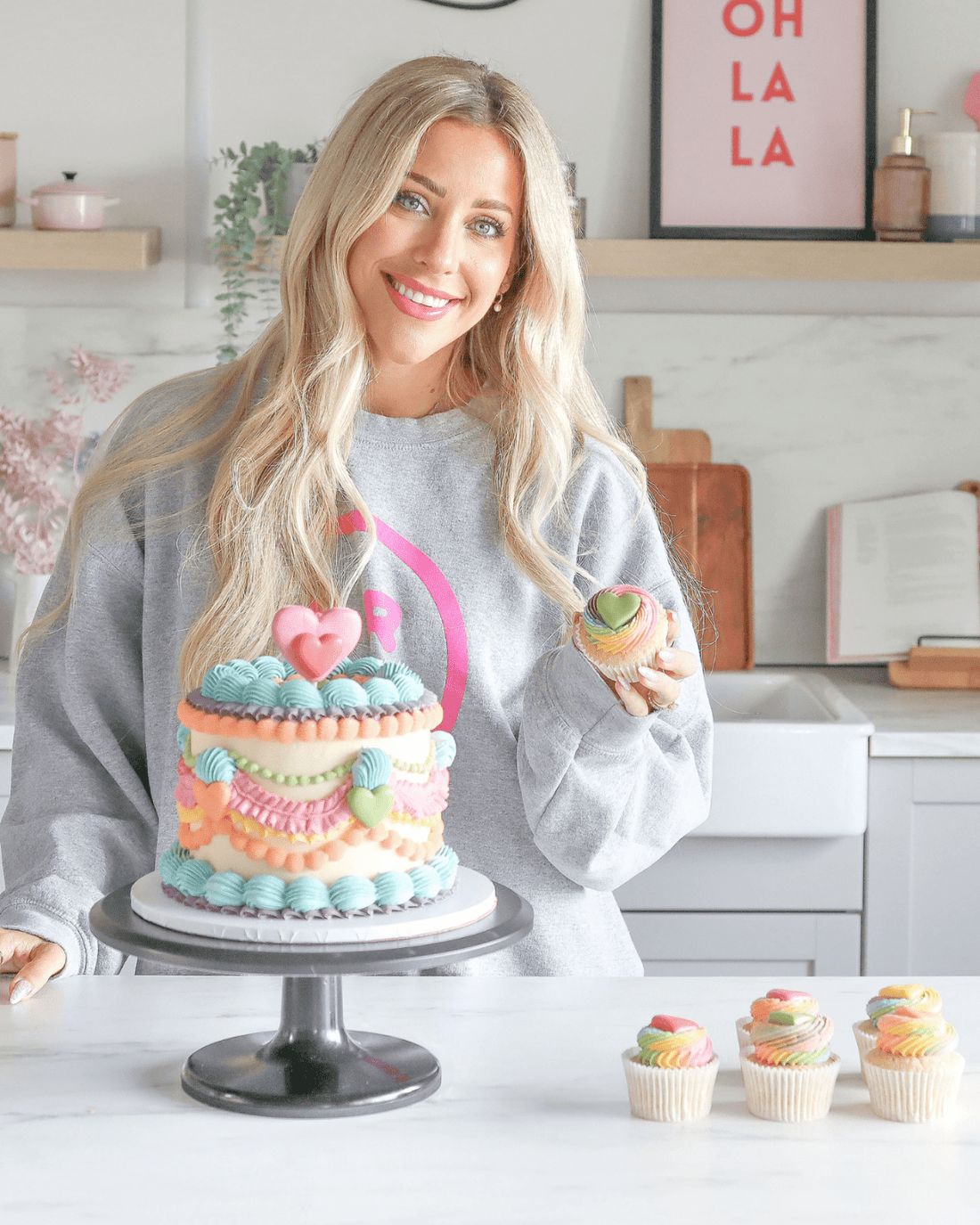 A smiling woman with long blonde hair stands in a kitchen holding a cupcake, with a Jenna Rae Cakes decorated cake and matching cupcakes, highlighting their Online Classes.