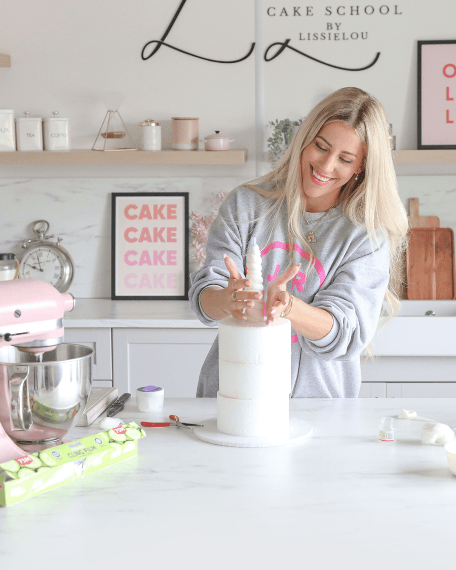 A woman smiles while decorating a tall white cake in a modern kitchen. Baking tools, a pink mixer, and cake supplies are on the counter, with Jenna Rae Cakes Online Classes visible on the wall behind her.