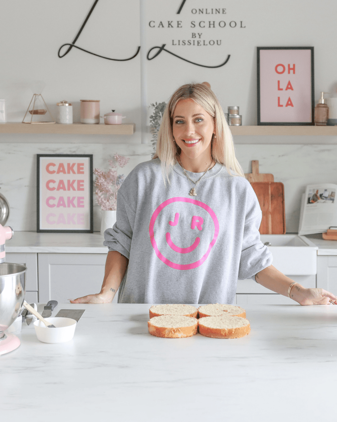 A smiling woman stands in a modern kitchen behind four slices of cake, wearing a gray sweatshirt with a pink smiley face and JR. The background features cake-themed decor and an OH LA LA sign, highlighting Jenna Rae Cakes Online Classes.