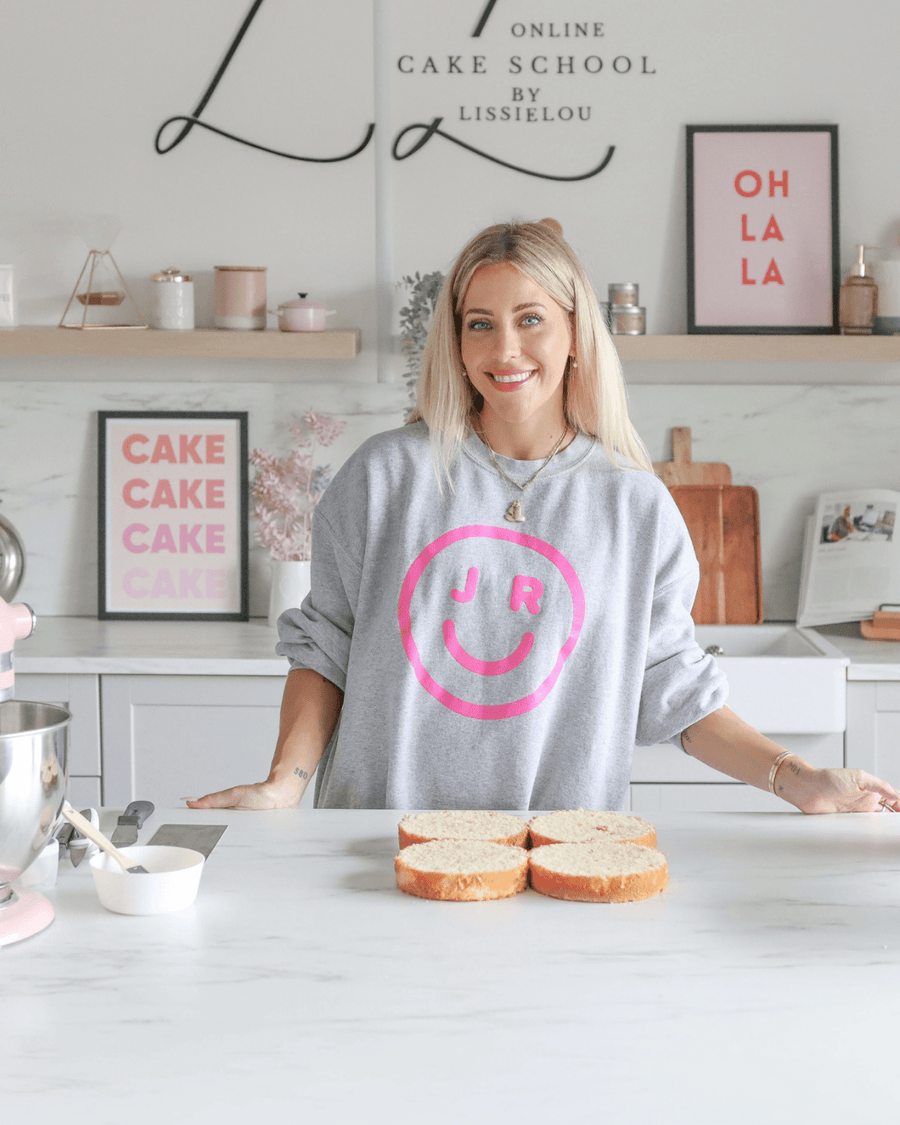 A smiling woman stands in a modern kitchen behind four slices of cake, wearing a gray sweatshirt with a pink smiley face and JR. The background features cake-themed decor and an OH LA LA sign, highlighting Jenna Rae Cakes Online Classes.
