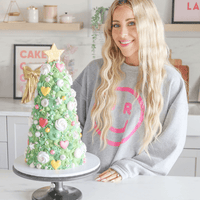 A smiling woman with long blonde hair and a gray sweatshirt stands behind a Christmas tree-shaped cake in a modern kitchen, highlighting Jenna Rae Cakes Online Classes.