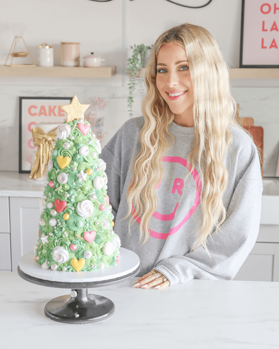 A smiling woman with long blonde hair and a gray sweatshirt stands behind a Christmas tree-shaped cake in a modern kitchen, highlighting Jenna Rae Cakes Online Classes.
