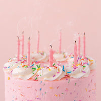 A Jenna Rae Cakes Party Candles set tops a pink frosted cake with colorful sprinkles and white icing swirls. Seven pink candles, just blown out and still smoking, stand against a pink background.