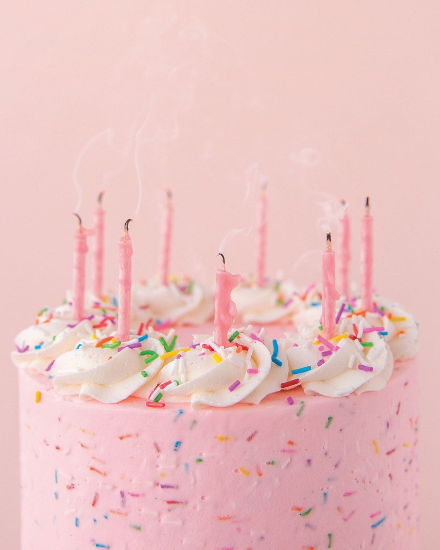 A Jenna Rae Cakes Party Candles set tops a pink frosted cake with colorful sprinkles and white icing swirls. Seven pink candles, just blown out and still smoking, stand against a pink background.