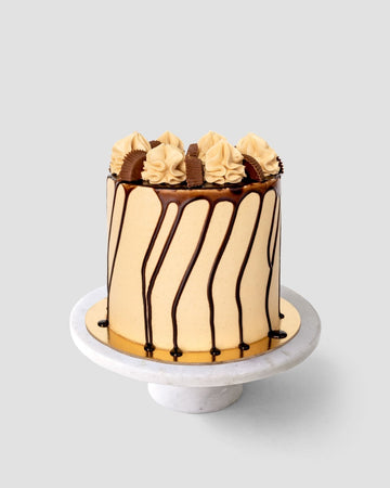 The Peanut Butter Cup Cake by Jenna Rae Cakes is a tall, round cake with light brown frosting, chocolate drip, and swirls of frosting and chocolate pieces on top, displayed on a white marble cake stand against a gray background.