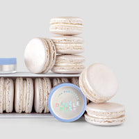 A stack of white macarons sits beside a box of more macarons and a round 5g shaker labeled Pearl Edible Lustre Dust by Jenna Rae Cakes, all arranged on a light gray background.
