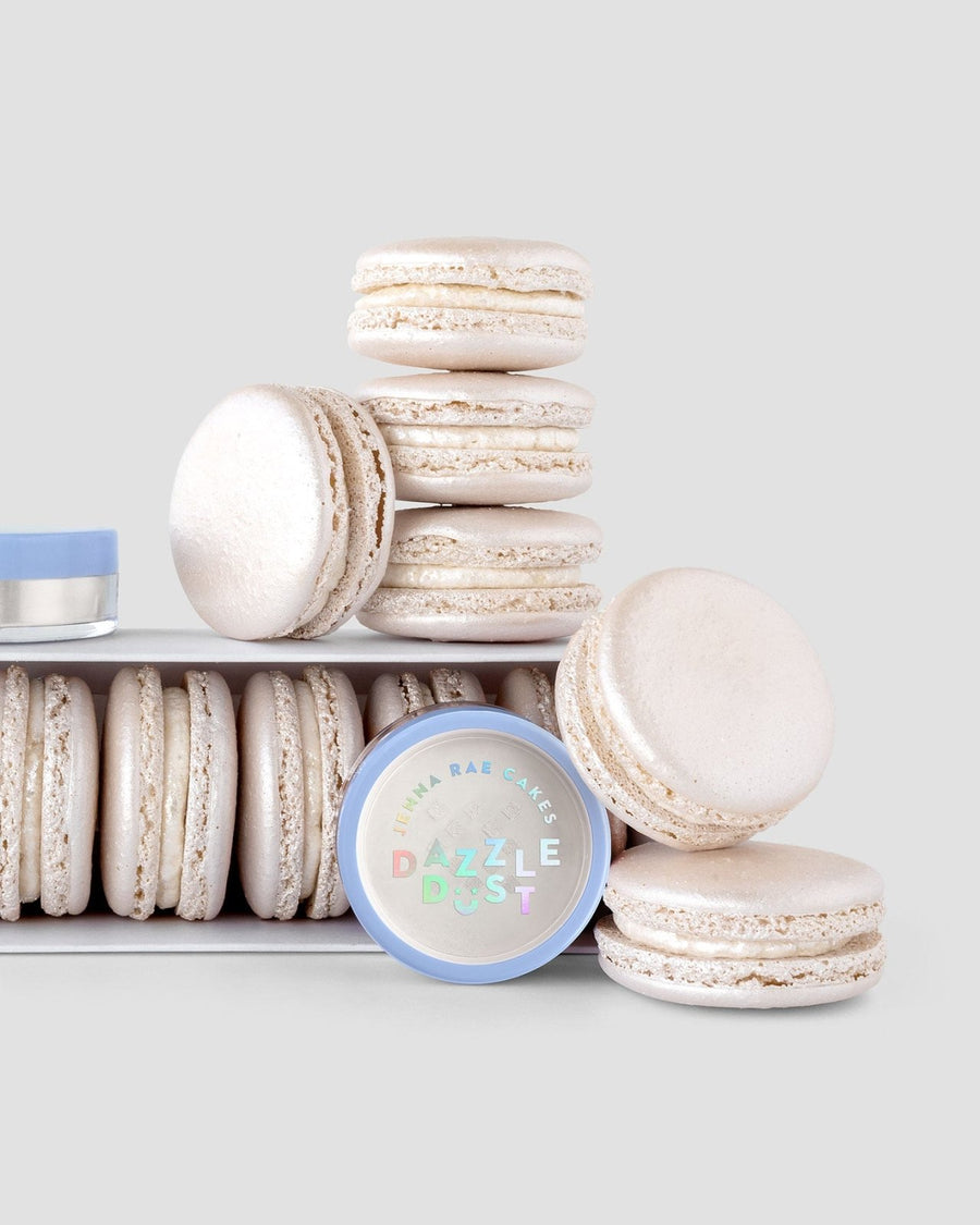 A stack of white macarons sits beside a box of more macarons and a round 5g shaker labeled Pearl Edible Lustre Dust by Jenna Rae Cakes, all arranged on a light gray background.