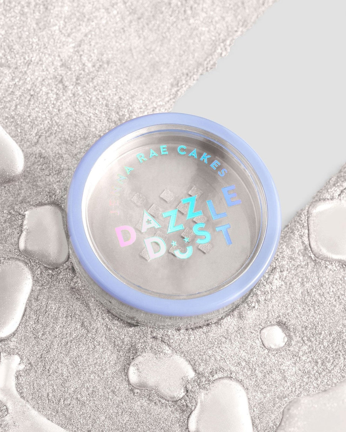 A round container of Jenna Rae Cakes Pearl Edible Lustre Dust (5g Shaker, Pack of 6) with a blue rim and holographic label rests on a silver textured surface, surrounded by droplets. The container holds shimmering edible powder.