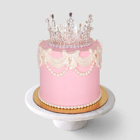 The Princess Crown Cake by Jenna Rae Cakes features pink frosting, white icing bows, pearly beads, and a decorative edible crown, presented on a round marble stand against a simple light background.