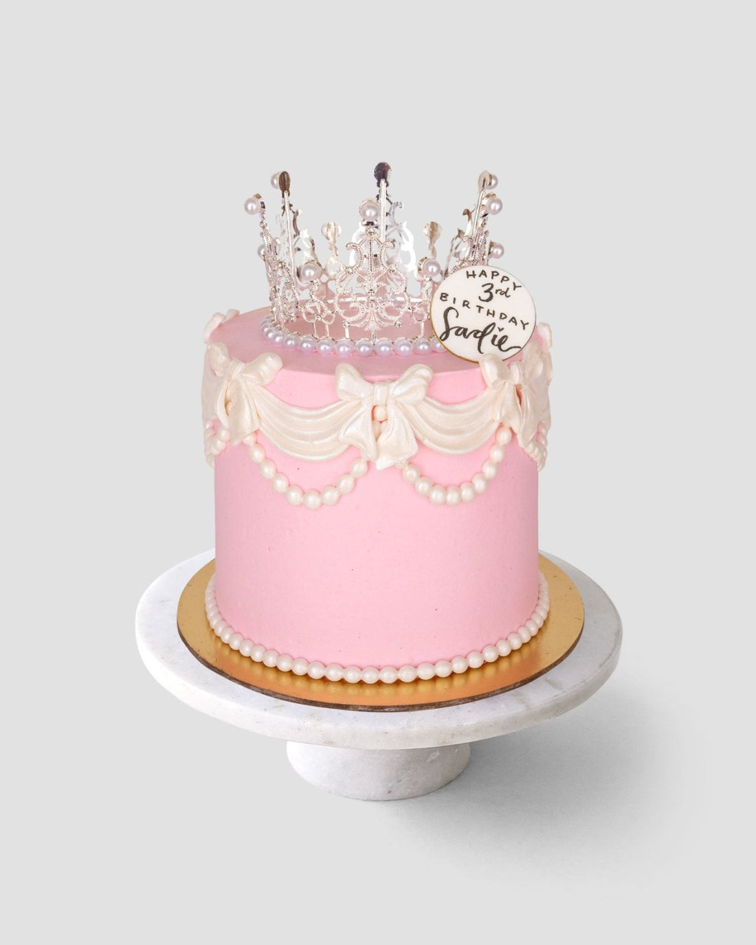 The Princess Crown Cake by Jenna Rae Cakes is a pink birthday cake with white icing bows, pearl decorations, a clear crown topper, and a plaque reading Happy 3rd Birthday, Charlie, displayed on a marble cake stand.