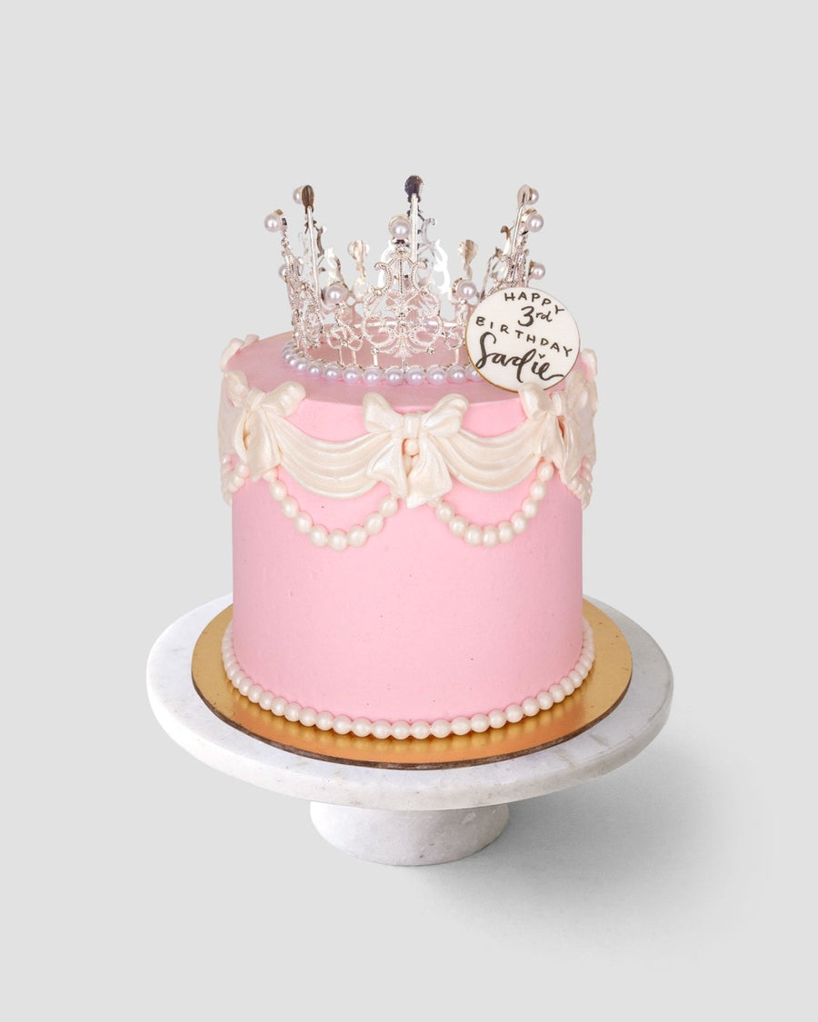 The Princess Crown Cake by Jenna Rae Cakes is a pink birthday cake with white icing bows, pearl decorations, a clear crown topper, and a plaque reading Happy 3rd Birthday, Charlie, displayed on a marble cake stand.
