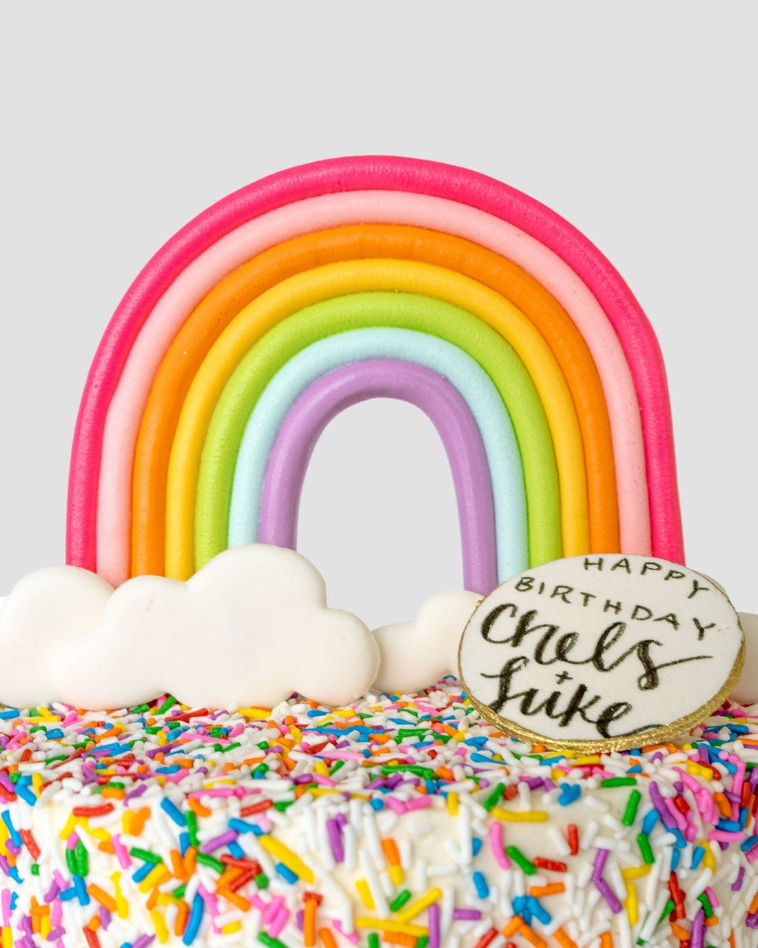 The Rainbow Cake by Jenna Rae Cakes is a colorful cake with rainbow and cloud decorations, rainbow sprinkles, and a round cookie on top that reads Happy Birthday Chels + Luke.