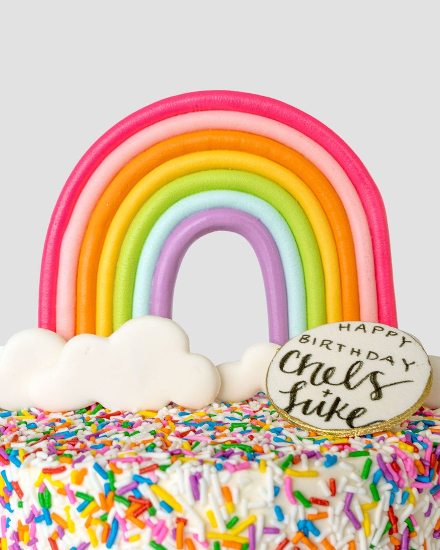 The Rainbow Cake by Jenna Rae Cakes is a colorful cake with rainbow and cloud decorations, rainbow sprinkles, and a round cookie on top that reads Happy Birthday Chels + Luke.