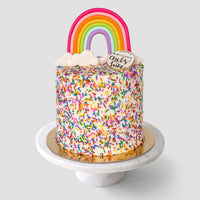 The Rainbow Cake by Jenna Rae Cakes is a tall, round cake covered in colorful sprinkles, topped with a fondant rainbow, clouds, and a small white plaque reading Happy chaos & cake, presented on a marble cake stand.