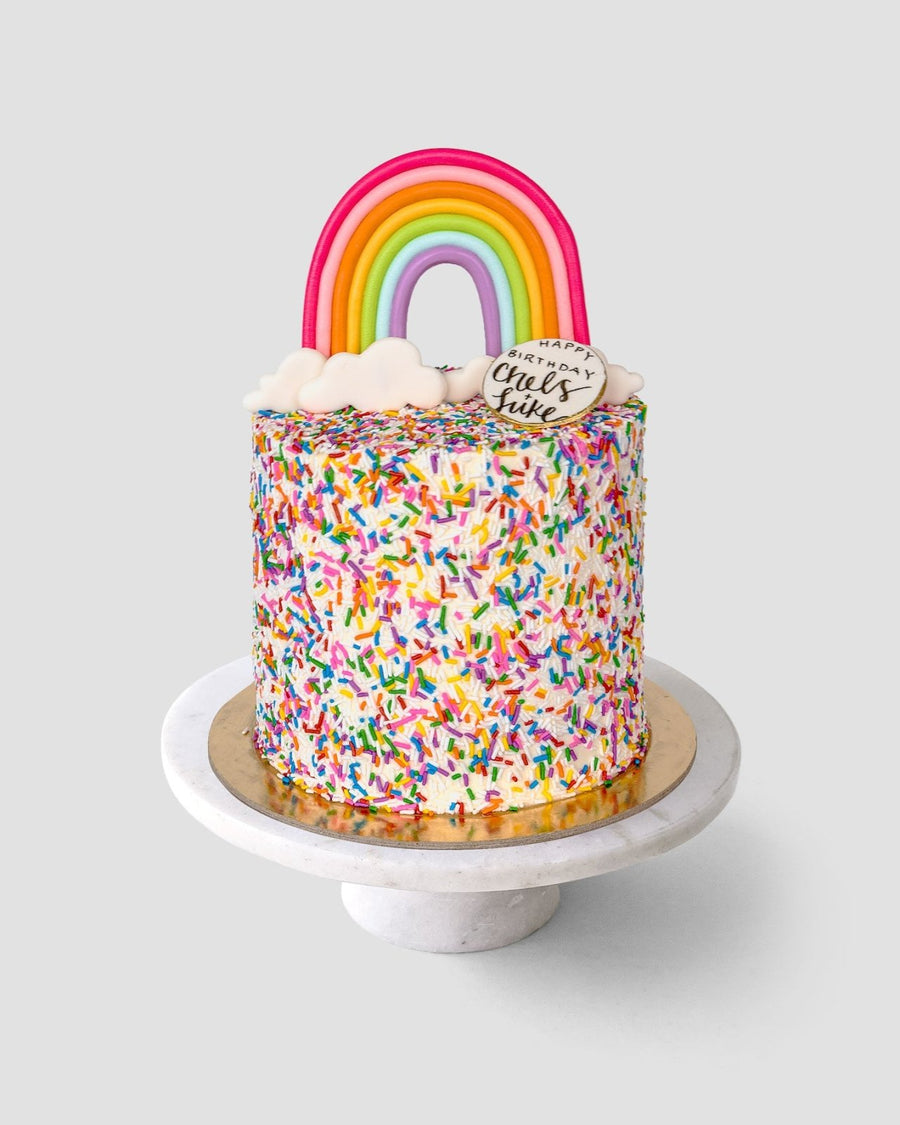 The Rainbow Cake by Jenna Rae Cakes is a tall, round cake covered in colorful sprinkles, topped with a fondant rainbow, clouds, and a small white plaque reading Happy chaos & cake, presented on a marble cake stand.