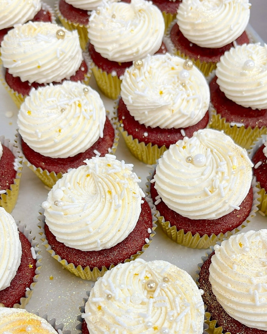 Rows of red velvet cupcakes with swirls of white frosting, topped with white and gold sprinkles, edible pearls, and glitter, arranged on a tray.