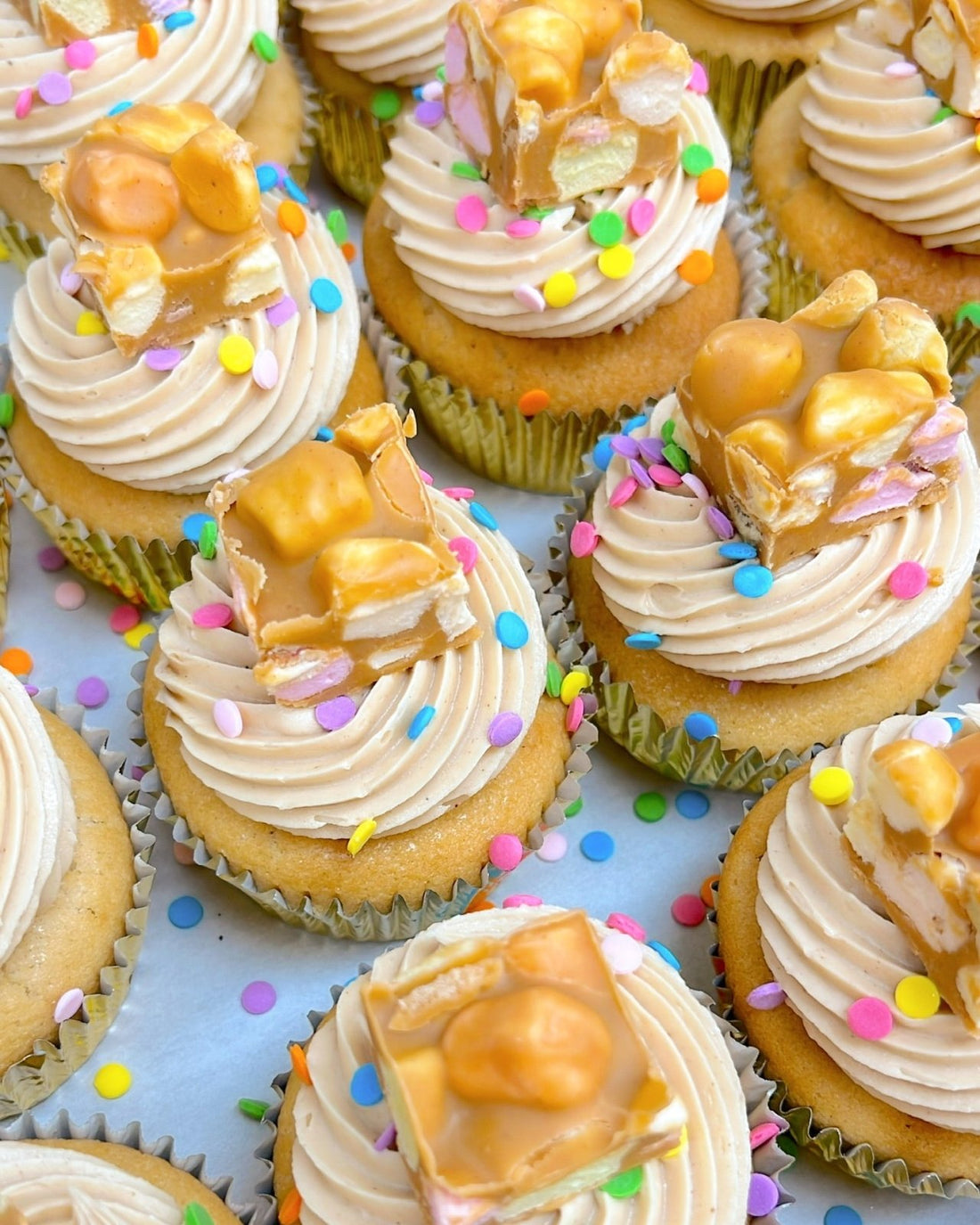 Regular Cupcakes by Jenna Rae Cakes are arranged closely together in gold wrappers and topped with swirls of frosting, colorful confetti sprinkles, and pieces of caramel peanut nougat candy on a white surface.