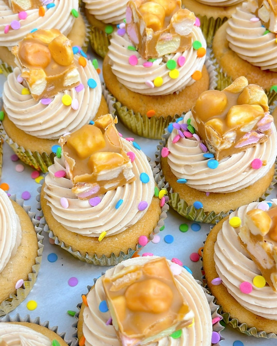 Regular Cupcakes by Jenna Rae Cakes are arranged closely together in gold wrappers and topped with swirls of frosting, colorful confetti sprinkles, and pieces of caramel peanut nougat candy on a white surface.