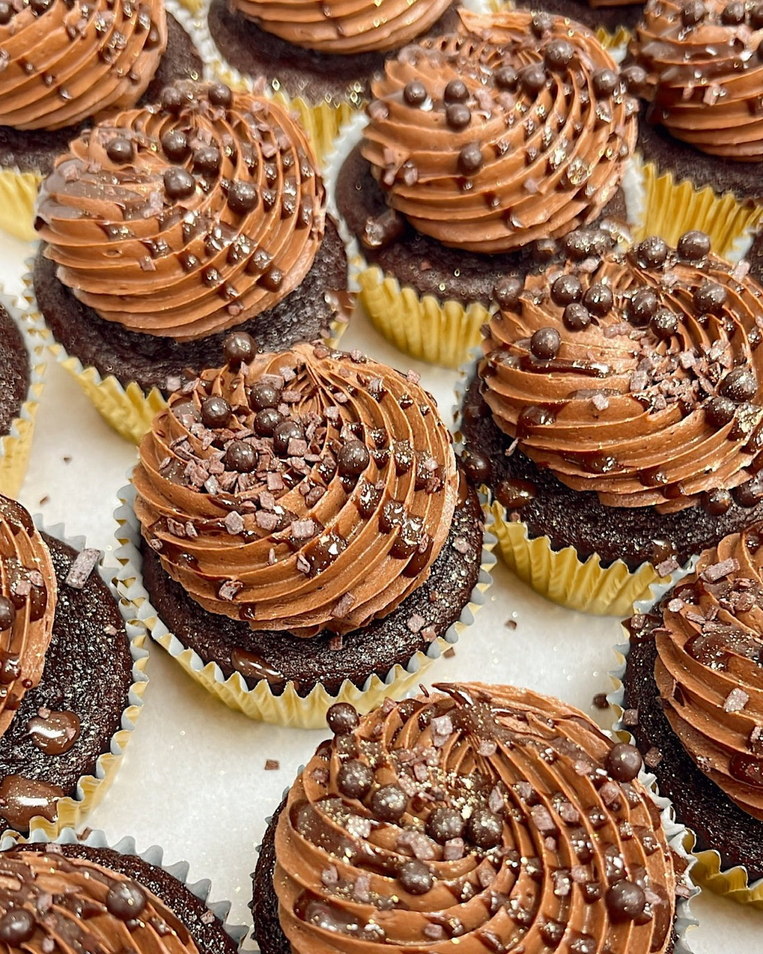 Chocolate cupcakes in yellow wrappers topped with swirls of chocolate frosting, chocolate sprinkles, and small chocolate pearls, arranged closely together on a white surface.