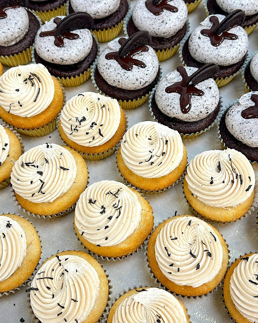 Rows of cupcakes on a tray; the front rows have yellow cake with swirls of creamy frosting topped with lavender, and the back rows have chocolate cupcakes with white frosting and a drizzle of chocolate.