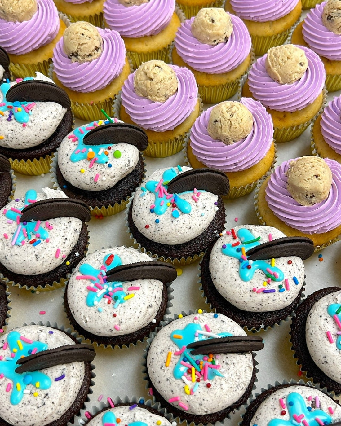 Rows of Jenna Rae Cakes Regular Cupcakes: chocolate with white frosting, blue drizzle, sprinkles, Oreo pieces, and vanilla with purple frosting topped with cookie dough balls.