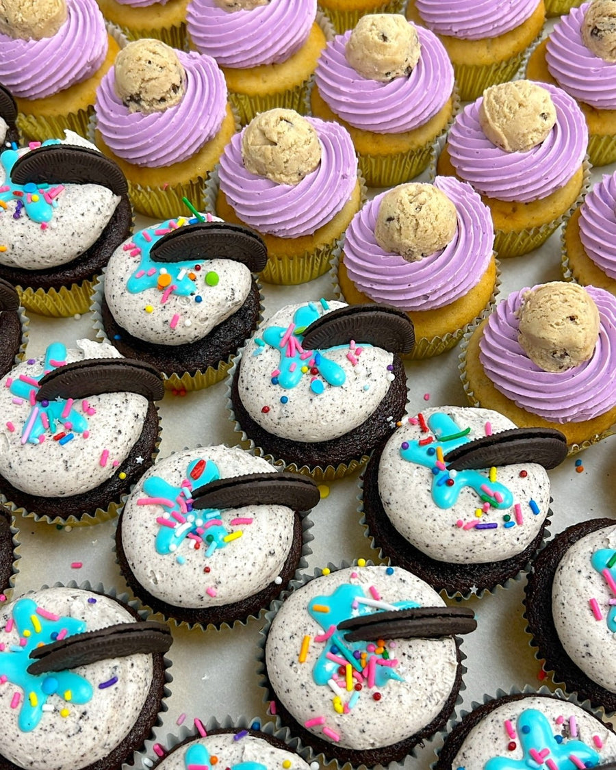 Rows of Jenna Rae Cakes Regular Cupcakes: chocolate with white frosting, blue drizzle, sprinkles, Oreo pieces, and vanilla with purple frosting topped with cookie dough balls.