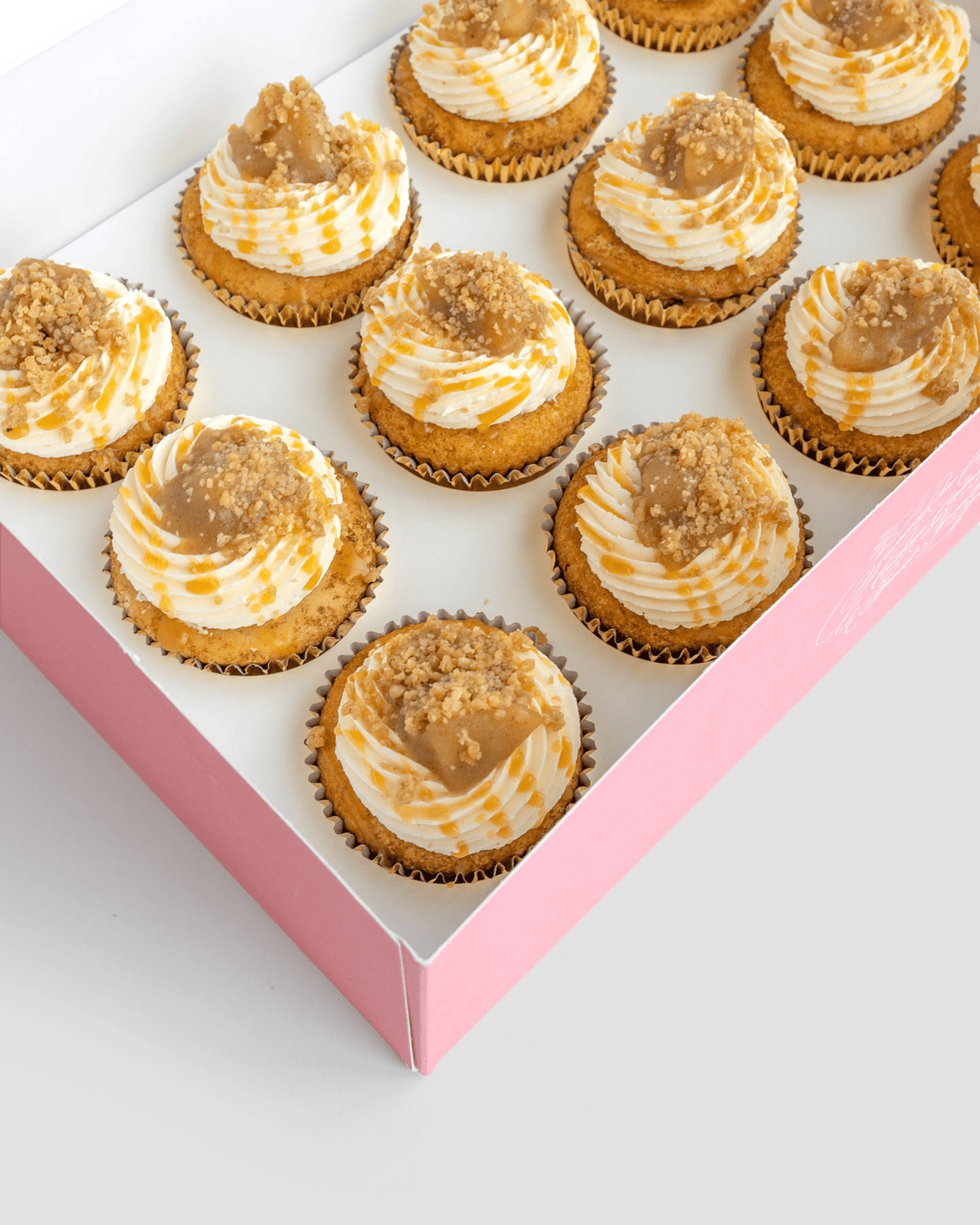 A pink box of twelve Regular Cupcakes by Jenna Rae Cakes, each topped with swirled white and caramel frosting and crumb topping, arranged neatly in rows.