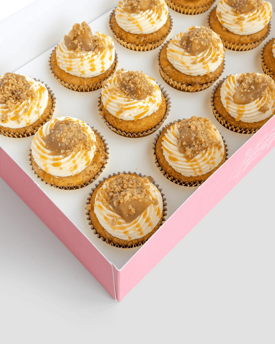 A pink box of twelve Regular Cupcakes by Jenna Rae Cakes, each topped with swirled white and caramel frosting and crumb topping, arranged neatly in rows.
