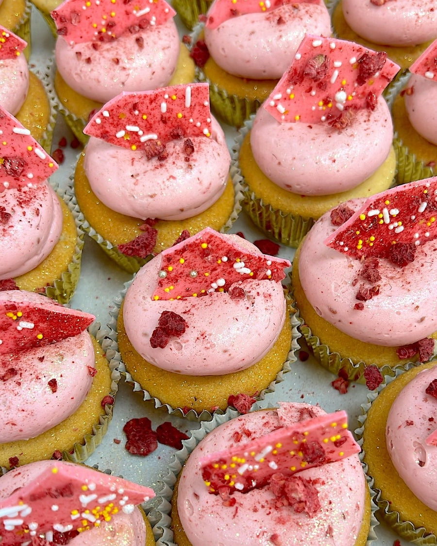 Regular Cupcakes by Jenna Rae Cakes are arranged in neat rows on a light background, each with a light yellow base, pink frosting, red candy shards, sprinkles, and crumbled red toppings.