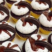 Chocolate cupcakes in yellow wrappers topped with cookies and cream frosting, a drizzle of chocolate sauce, and a piece of chocolate sandwich cookie on each cupcake, arranged closely together in rows.
