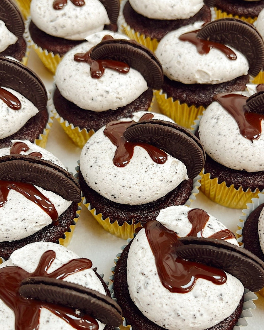 Chocolate cupcakes in yellow wrappers topped with cookies and cream frosting, a drizzle of chocolate sauce, and a piece of chocolate sandwich cookie on each cupcake, arranged closely together in rows.