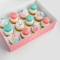 The Reveal Works Cupcakes by Jenna Rae Cakes come in a pink box with cupcakes topped with blue and pink macarons, pastel meringue swirls, and a white fondant cupcake reading He or she?—perfect for a gender reveal celebration.
