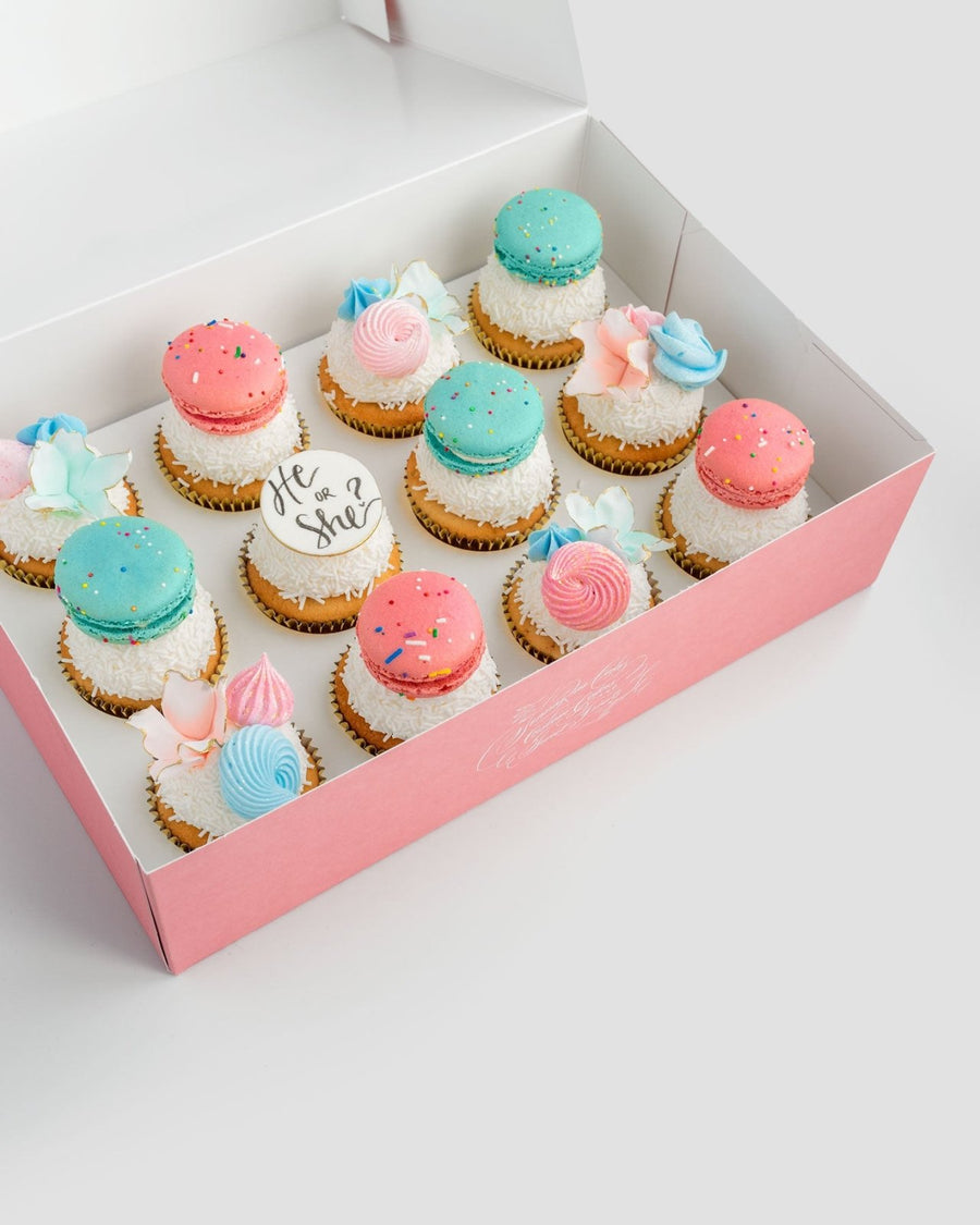 The Reveal Works Cupcakes by Jenna Rae Cakes come in a pink box with cupcakes topped with blue and pink macarons, pastel meringue swirls, and a white fondant cupcake reading He or she?—perfect for a gender reveal celebration.