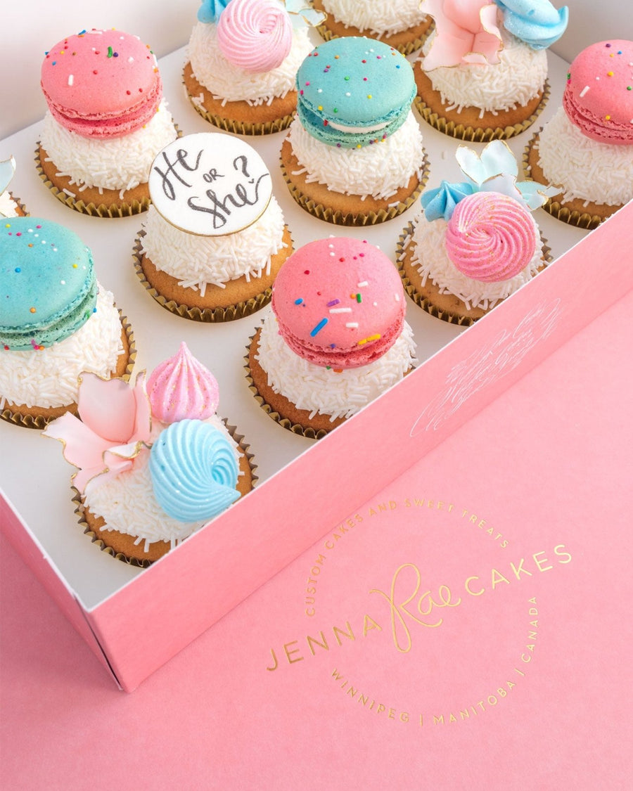 A pink box of Jenna Rae Cakes Reveal Works Cupcakes, decorated with pink and blue macarons (one reads He or She?), pastel meringues, and white sprinkles.