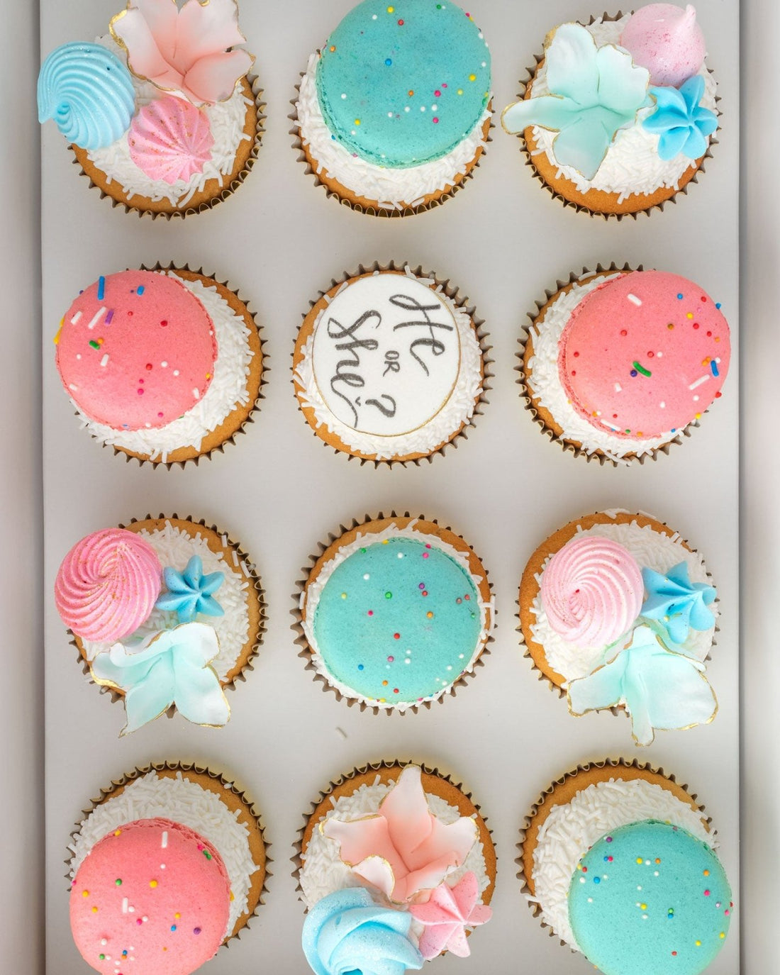The Reveal Works Cupcakes by Jenna Rae Cakes feature 12 cupcakes topped with pink and blue macarons, pastel flowers, sprinkles, and a center cupcake reading “He or She?” for a charming gender reveal.