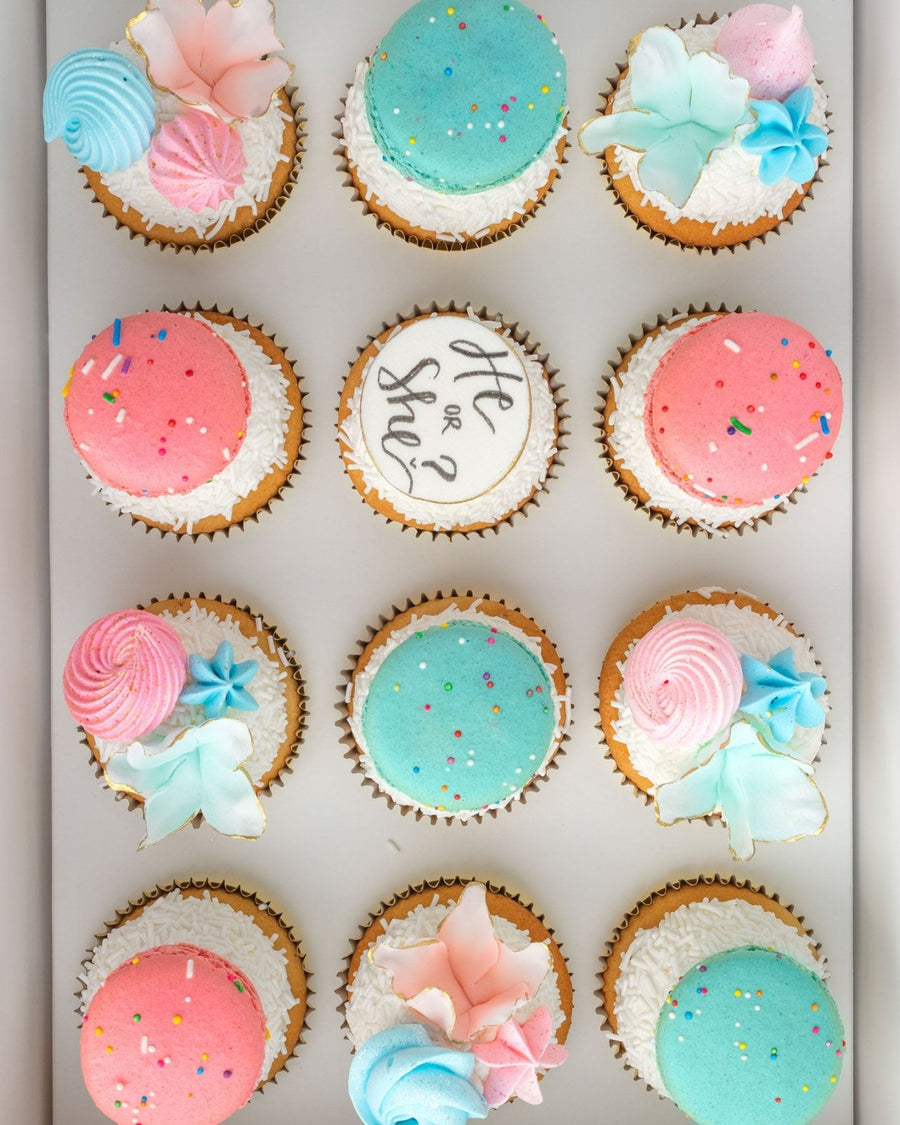 The Reveal Works Cupcakes by Jenna Rae Cakes feature 12 cupcakes topped with pink and blue macarons, pastel flowers, sprinkles, and a center cupcake reading “He or She?” for a charming gender reveal.