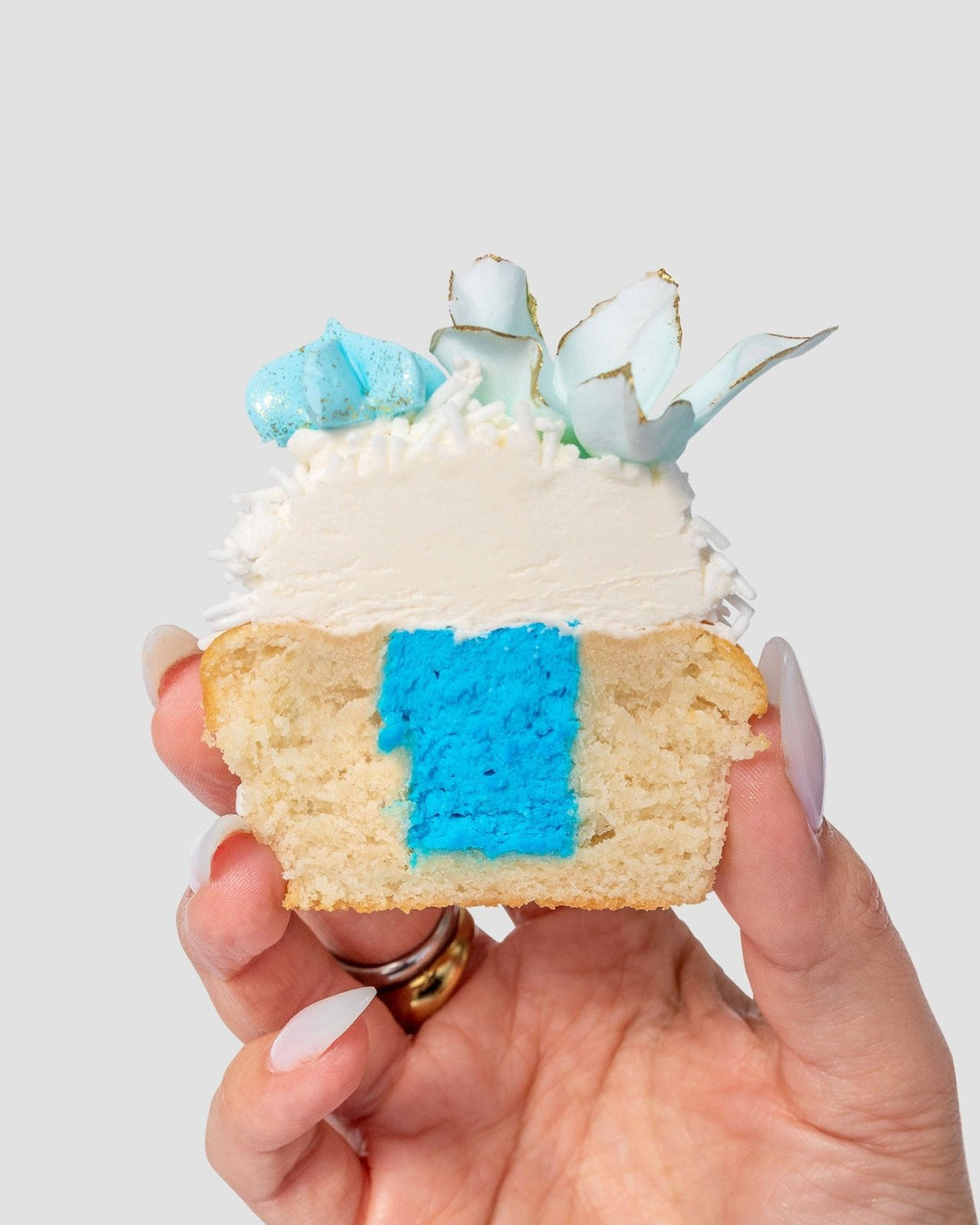 A hand holds half of a Reveal Works Cupcake by Jenna Rae Cakes, topped with white and blue icing, shredded coconut, and a decorative blue and white accent, revealing a bright blue filling inside.