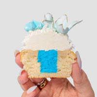 A hand holds half of a Reveal Works Cupcake by Jenna Rae Cakes, topped with white and blue icing, shredded coconut, and a decorative blue and white accent, revealing a bright blue filling inside.
