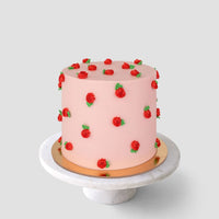 The Rose Bud Cake by Jenna Rae Cakes is a round, pink frosted cake topped with small red buttercream roses and green leaves, shown on a white marble stand against a plain light gray background.