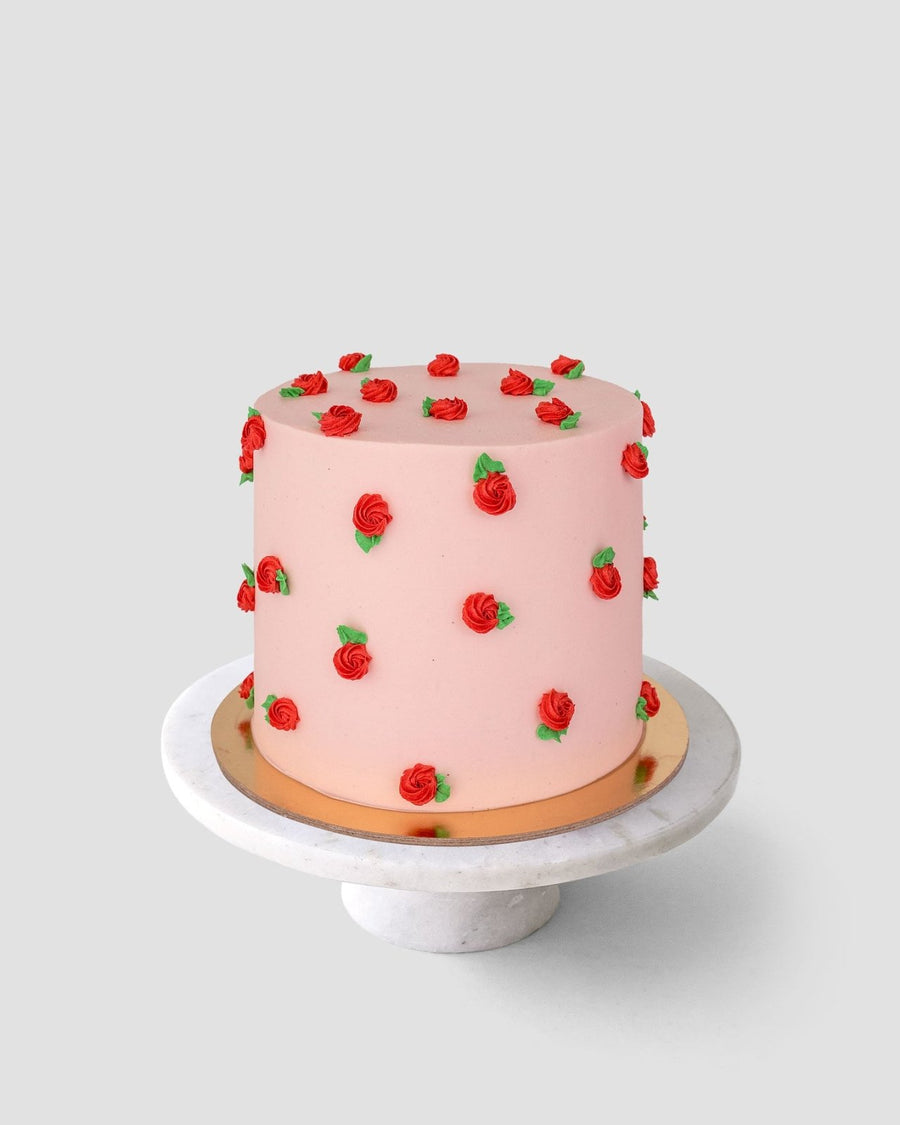 The Rose Bud Cake by Jenna Rae Cakes is a round, pink frosted cake topped with small red buttercream roses and green leaves, shown on a white marble stand against a plain light gray background.