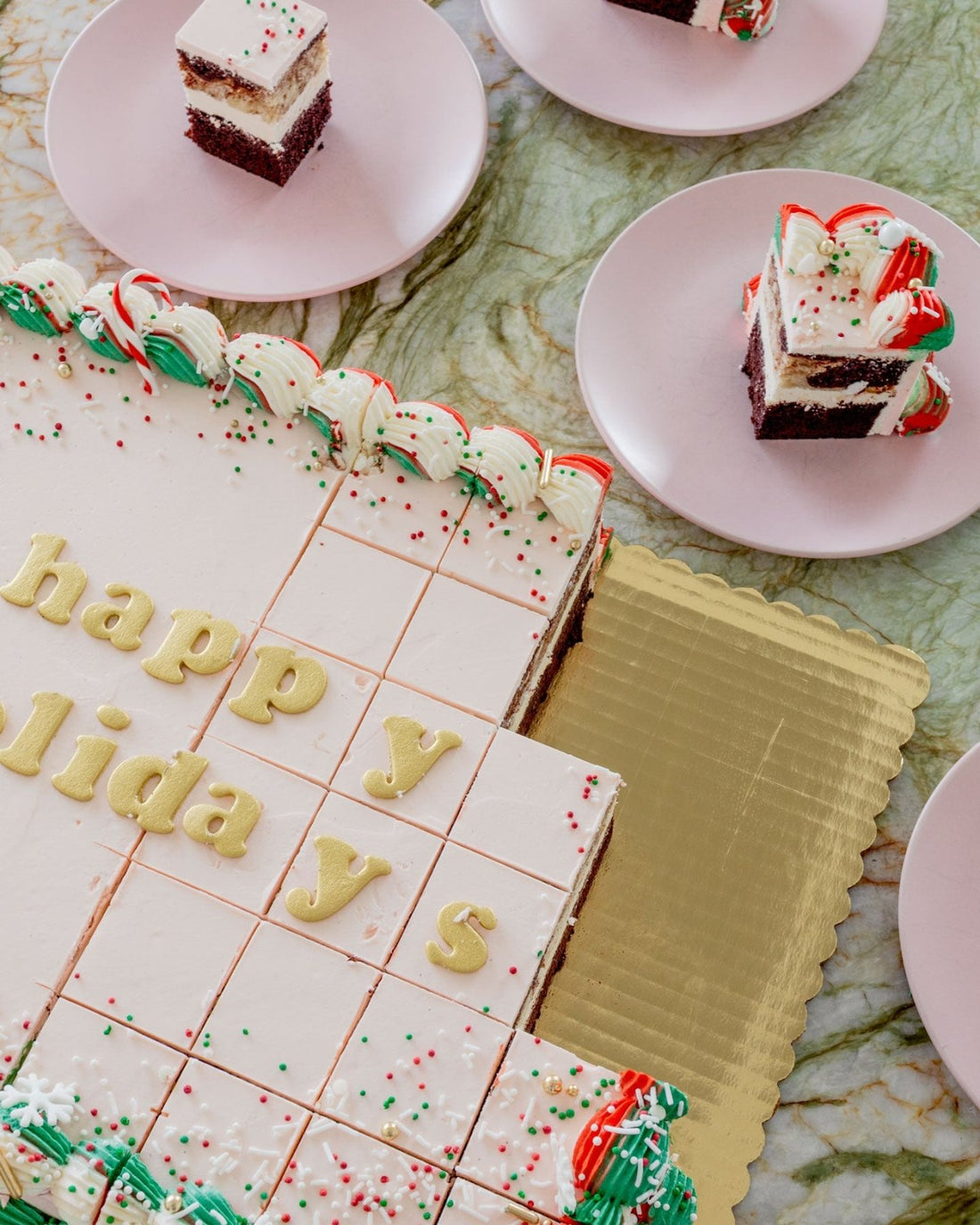 The Sugar Flower Sheet Cake by Jenna Rae Cakes, a large rectangular cake with white icing and colorful sprinkles, sits on a gold tray. Happy Holidays is written on top, and slices are served on pink plates around it.