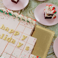 The Sugar Flower Sheet Cake by Jenna Rae Cakes, a large rectangular cake with white icing and colorful sprinkles, sits on a gold tray. Happy Holidays is written on top, and slices are served on pink plates around it.