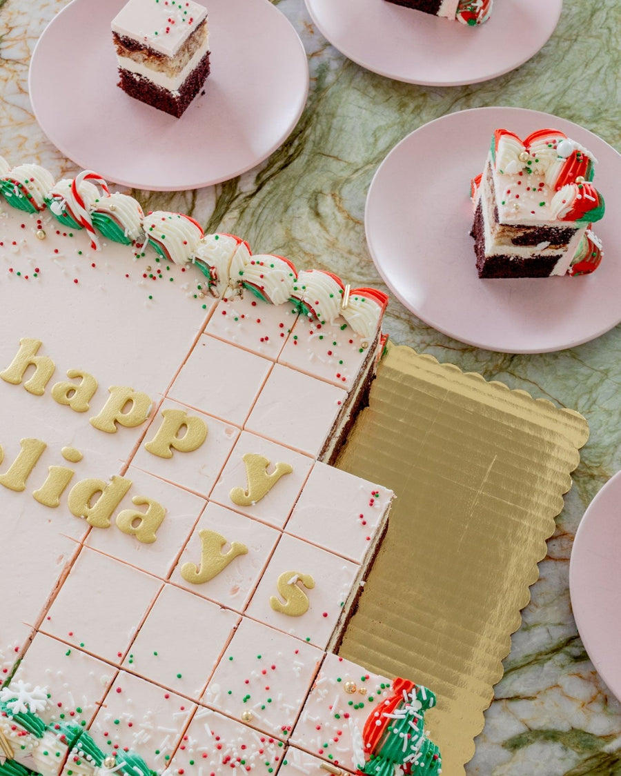 The Sugar Flower Sheet Cake by Jenna Rae Cakes, a large rectangular cake with white icing and colorful sprinkles, sits on a gold tray. Happy Holidays is written on top, and slices are served on pink plates around it.