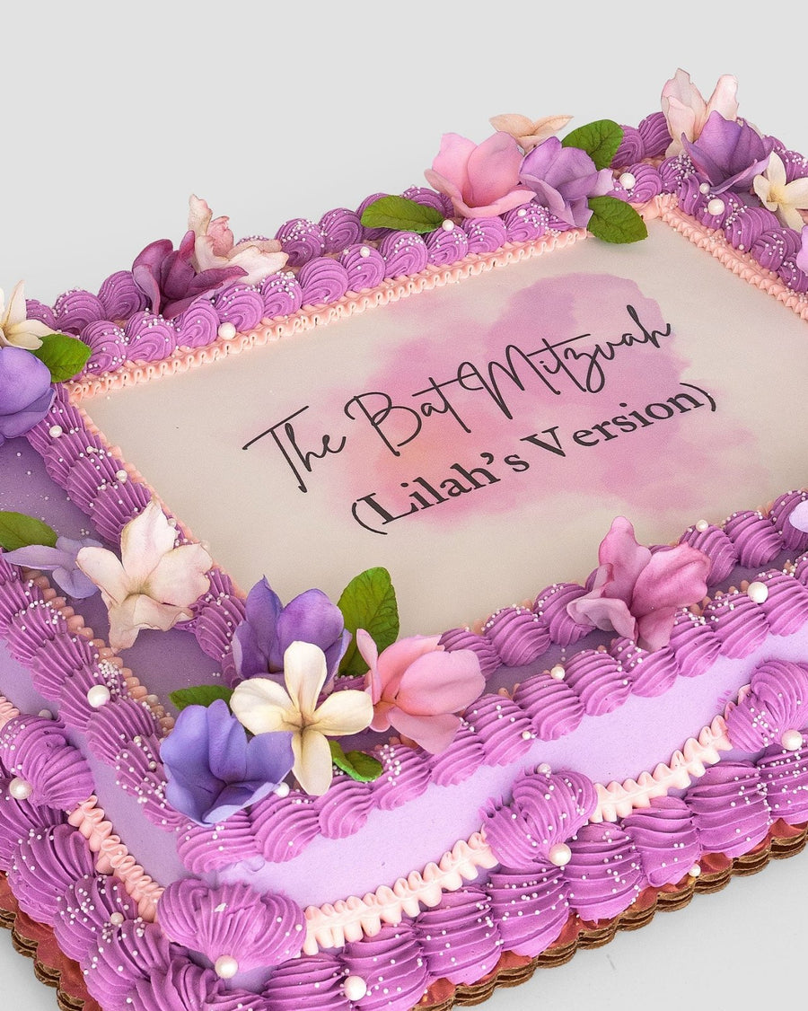 The Sugar Flower Sheet Cake by Jenna Rae Cakes is a rectangular cake with purple and pink frosting, sugar flowers, green leaves, and The Bat Mitzvah (Lilah’s Version) written in elegant black script on a white center panel.